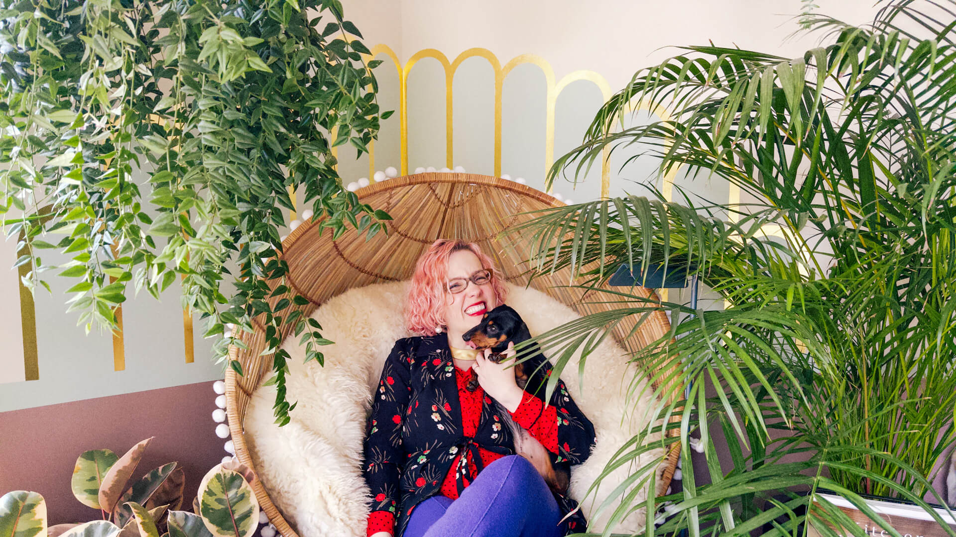 Alexandria sitting with Remi (the mini dachshund) on her living room plant throne. Alexandria is smiling, wearing brightly colour clothing and Remi is looking cute—he just can't help it.